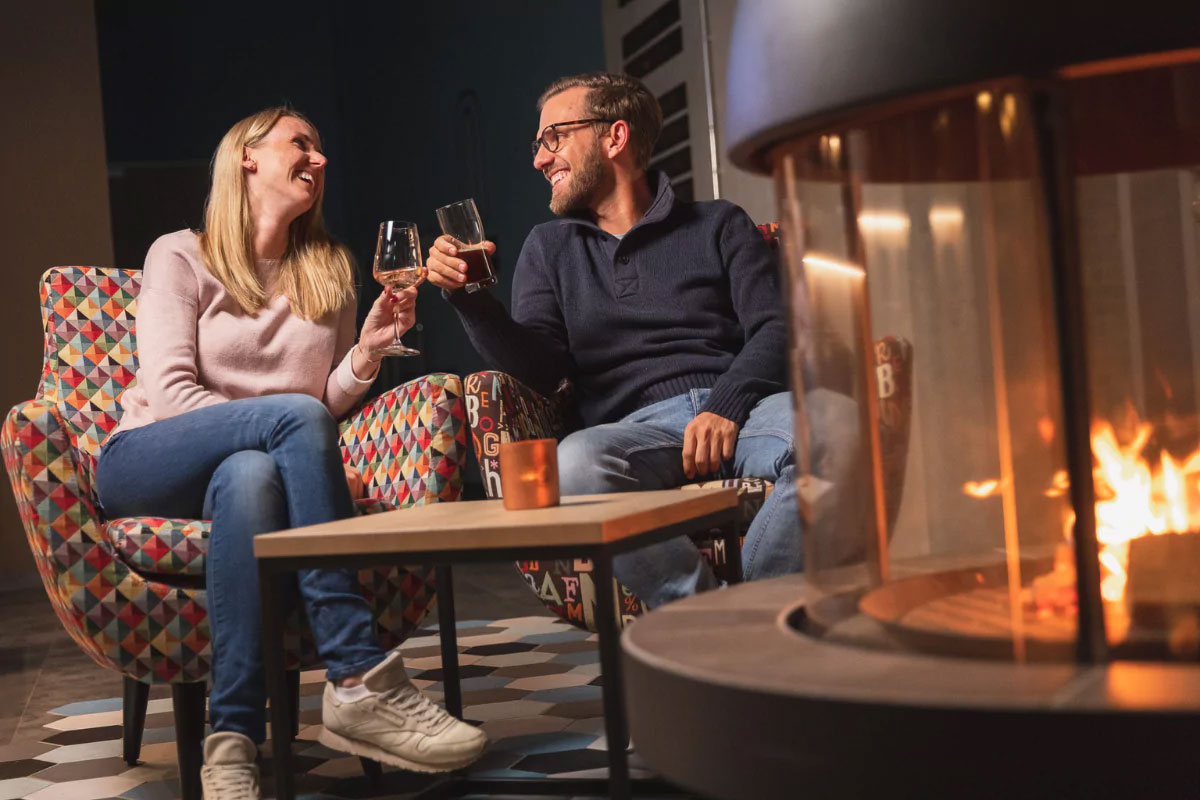 Ein Pärchen sitzt an einem modernen Kamin und stößt mit einem Glas mit Getränk an. Die blonde Frau lacht, der Mann mit der Brille lächelt sie an.