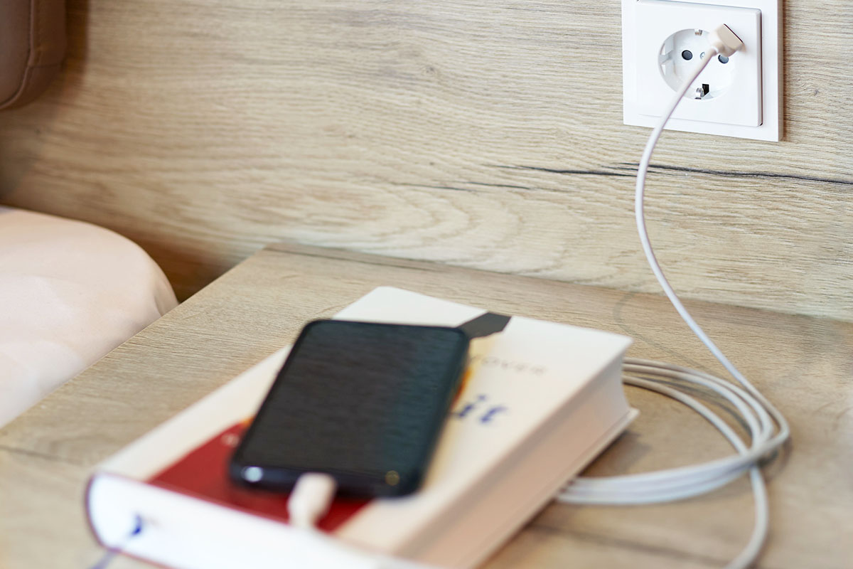 Ein Handy liegt auf dem Beistelltisch neben dem Bett in der ancora Suite im arborea Marina Resort auf einem Buch und wird an der Steckdose aufgeladen.