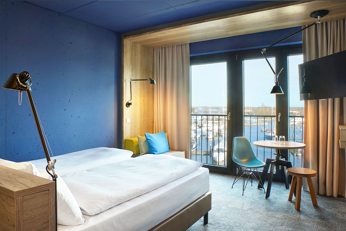 Kingsize Doppelbett, Sitzgelegenheit, Tisch mit Stühlen, TV und Blick auf die Neustädter Bucht im Doppelzimmer Marina im arborea Marina Resort Neustadt.