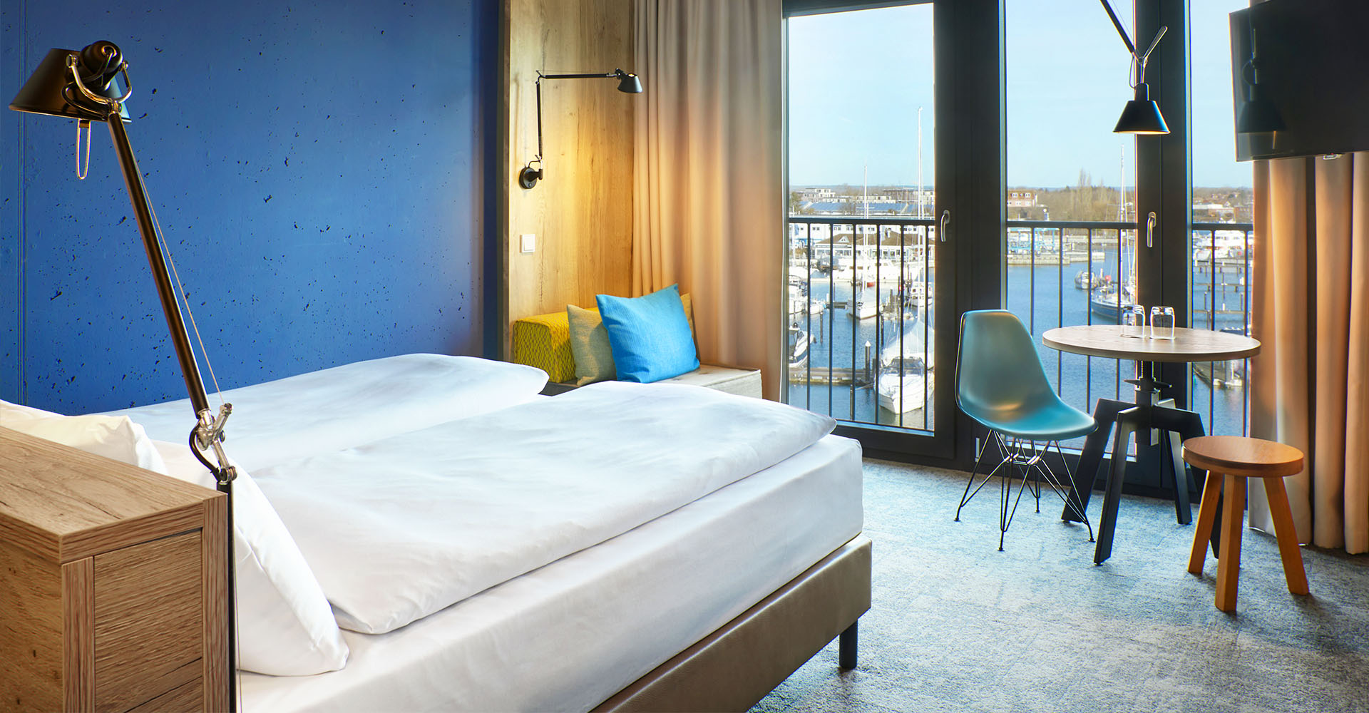 Kingsize Doppelbett mit Sitzgelegenheit, Esstisch mit Stühlen, TV und einem Ausblick auf die Neustädter Bucht im Doppelzimmer Marina im arborea Marina Resort Neustadt.