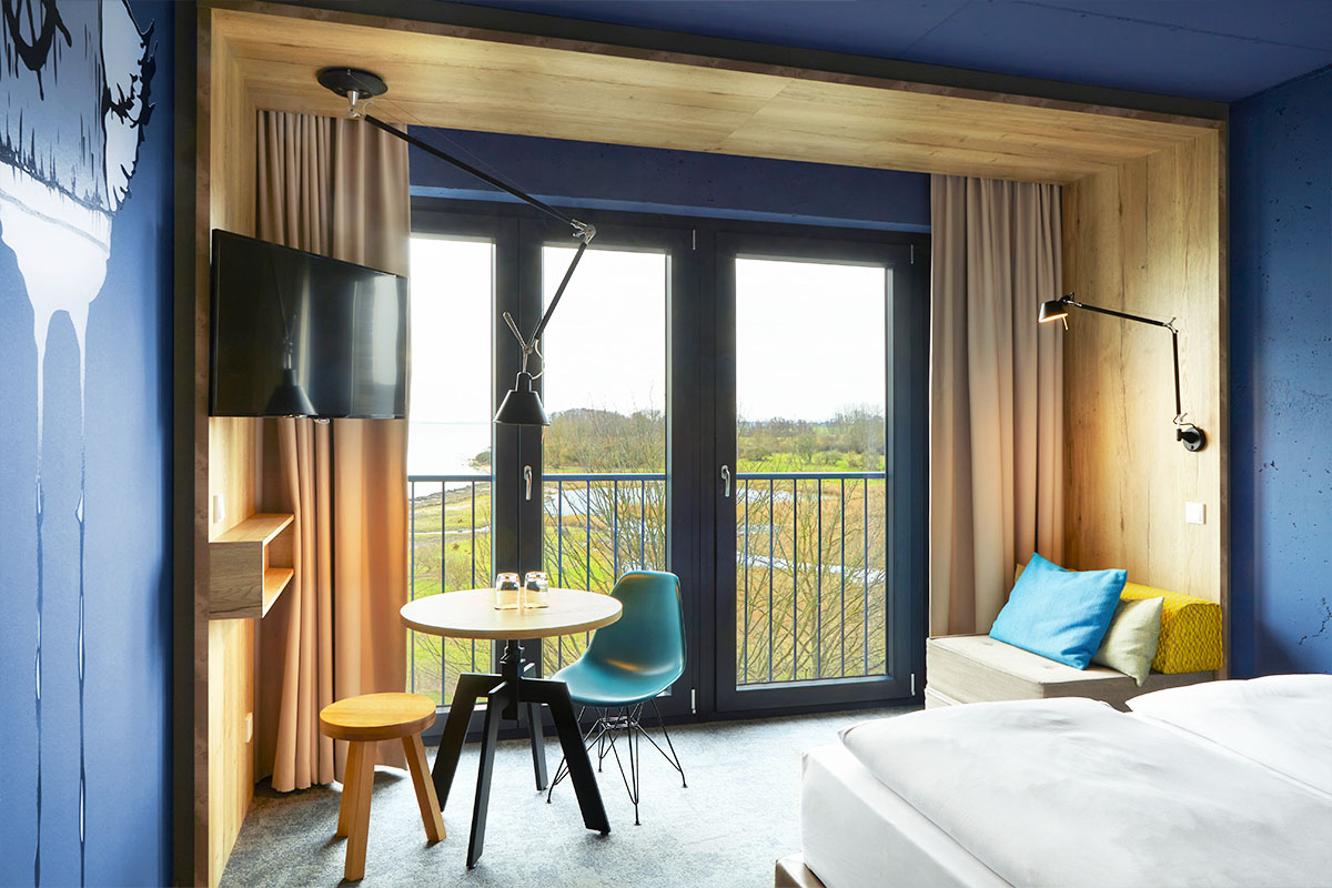 Die Inneneinrichtung des Doppelzimmer Ostsee im arborea Marina Resort Neustadt mit einem TV, einem Essbereich mit Tisch und zwei Stühlen, einem kleinen Sessel und einem Kingsize Bett mit Landblick.